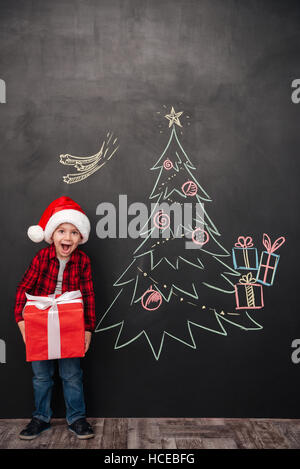 Bild der fröhlichen schreiendes Kind Hut hält ein großes Geschenk Weihnachtsbaum auf Tafel zeichnen. Blick in die Kamera. Stockfoto