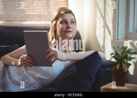 Nachdenkliche Frau liegt auf Sofa und hält digitale Tisch im Wohnzimmer Stockfoto