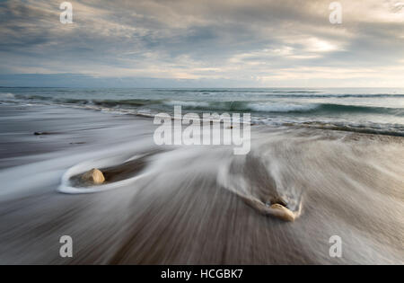 Kieselsteine am Strand auf einem schwarzen Sand und Meer Wasser fließt bei Sonnenuntergang Erstellen schöne Texturen. lange Belichtung Bild. Stockfoto