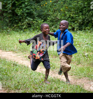 Zwei Jungen aus Sierra Leone sind glücklich und laufen durch ihr ländliches Dorf. Das T-Shirt zaudert: 'Die Reichen weinen auch'. Zwei arme Jungen laufen und genießen das Leben. In der Gola-Gegend leben Menschen aus dem Stamm Mendy. Ein Junge trägt seine Schuluniform, der andere hat Stifte in der Hand und trägt ein T-Shirt, das ihm wahrscheinlich von Reisenden geschenkt wurde. Stockfoto