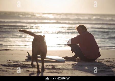 Mann mit Surfbrett sitzt mit seinem Hund am Strand Stockfoto