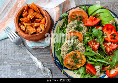 Rucola, vegane Linsensuppe gebratene Schnitzel, gebackene Süßkartoffeln, Tomaten, Avocado, Pfeffer. Gebackene Süßkartoffel mit Kräutern der Provence in einem Topf auf einem Woode Scheiben Stockfoto