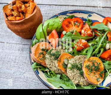 Rucola, vegane Linsensuppe gebratene Schnitzel, gebackene Süßkartoffeln, Tomaten, Avocado, Pfeffer. Gebackene Süßkartoffel mit Kräutern der Provence in einem Topf auf einem Woode Scheiben Stockfoto