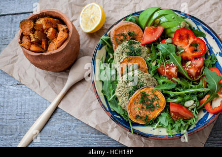 Rucola, vegane Linsensuppe gebratene Schnitzel, gebackene Süßkartoffeln, Tomaten, Avocado, Pfeffer. Gebackene Süßkartoffel mit Kräutern der Provence in einem Topf auf einem Woode Scheiben Stockfoto