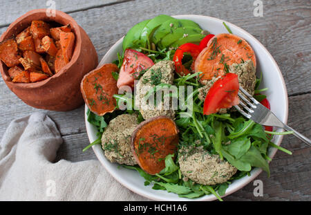 Rucola, vegane Linsensuppe gebratene Schnitzel, gebackene Süßkartoffeln, Tomaten, Avocado, Pfeffer. Gebackene Süßkartoffel mit Kräutern der Provence in einem Topf auf einem Woode Scheiben Stockfoto