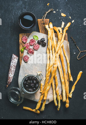 Wein und Häppchen festgelegt. Italienische Grissini Brot Sticks, trockenen Wurstwaren Fleischwurst, schwarzen Oliven im Glas und Rotwein im Glas auf hölzernen Portion board Stockfoto