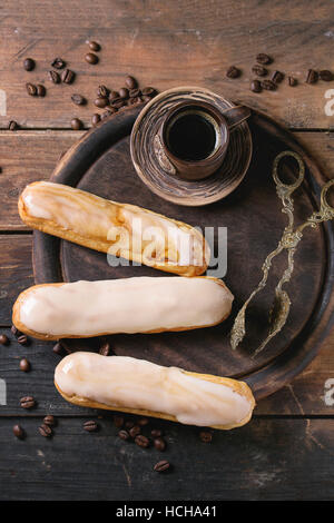 Frische hausgemachte Eclairs mit verschiedenen Glasur, Kaffeebohnen und Keramik Tasse schwarzen Kaffee über alte Holz Textur Hintergrund mit Platz für Text. Nach oben Stockfoto