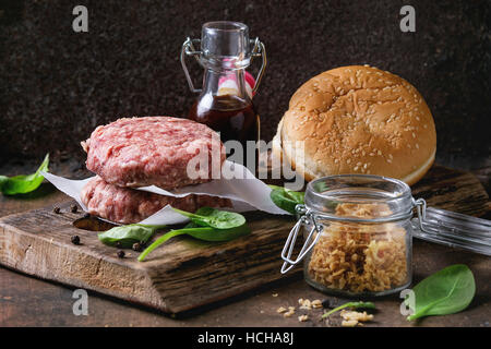 Zutaten für die Herstellung der Hamburger. Rohe Burger Rind Schnitzel, zwei Brötchen, gebratenen Zwiebeln, frischem Spinat und Ketchup-sauce auf dunklem Holz Schneidebrett über alte Stockfoto