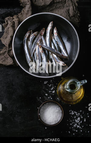 Viele Fische roh frische Sardellen in schwarzen Keramikschale mit Meersalz und Flasche Olivenöl Marinade über schwarzen Metall Hintergrund. Ansicht von oben. Meer Stockfoto