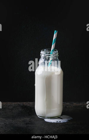 Geöffnete Glasflasche voll Milch mit Spot von Milch und gestreiften retro cocktail Rohr auf schwarzem Hintergrund. Textfreiraum Stockfoto