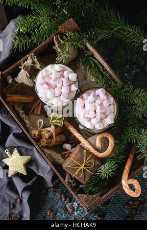 Zwei Gläser Café Latte mit rosa Marshmallow stehend in Holzkiste mit Dekor, Bonbons, Gewürze, Kaffee und Tanne Weihnachtsbaum über dunkle Textur ba Stockfoto