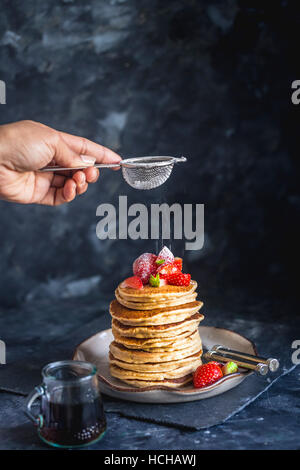 Abstauben Icing Sugar auf Stapel von Gluten freie Hafer Mehl Pfannkuchen serviert mit gehackten Erdbeeren und Ahornsirup Stockfoto