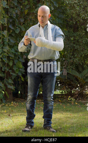 Junger Mann mit einem Arm und Ellenbogen in einem weißen Putz / Fiberglas steht in einem Garten SMS auf seinem Handy