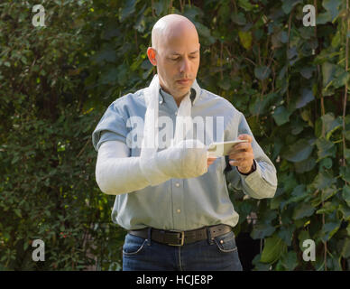 Junger Mann mit einem Arm und Ellenbogen in einem weißen Putz / Fiberglas steht in einem Garten SMS auf seinem Handy