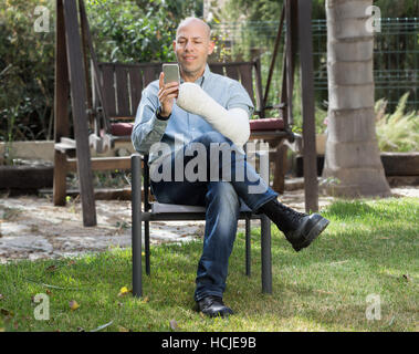 Junger Mann mit einem Arm und Ellenbogen in einem weißen Putz / Fiberglas sitzen in einem Garten SMS auf seinem Handy