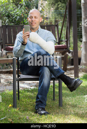 Junger Mann mit einem Arm und Ellenbogen in einem weißen Putz / Fiberglas sitzen in einem Garten SMS auf seinem Handy
