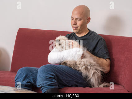 Junger Mann mit einem Arm und Ellenbogen in einem weißen Putz / Fiberglas gegossen zu Hause entspannen nach den Arm gebrochen hatte seine Katze streicheln Stockfoto