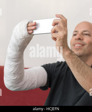 Junger Mann mit einem Arm und Ellenbogen in einem weißen Putz / Fiberglas gegossen zu Hause sprechen ein Selbstporträt auf seinem Smartphone während der Genesung von einem gebrochenen Arm Stockfoto