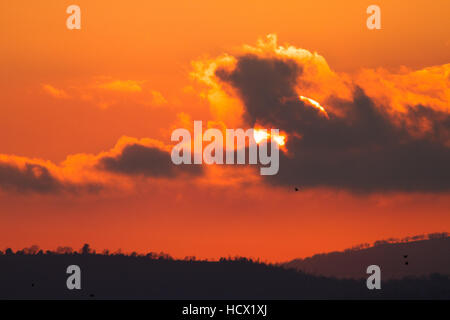 Landschaft mit roten Sonnenuntergang hinter dunklen Wolken und Silhouette von Hügeln und Bäumen Stockfoto