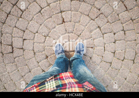 Männliche Füße in Blue Jeans und Turnschuhe stehen auf alten grauen Steinpflaster mit runden Muster Stockfoto