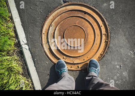 Männliche Füße in blauen Sportschuh auf Straßenpflaster in der Nähe von Runde verrosteten Kanalisation Kanaldeckel Stockfoto