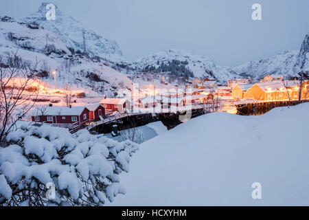 Dämmerung leuchtet das Fischerdorf umgeben von schneebedeckten Gipfeln, Nusfjord, Nordland, Lofoten-Inseln, Nord-Norwegen Stockfoto