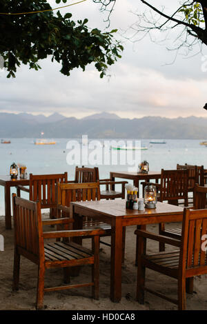 Leere Holztischen & Stühle in einem Strandrestaurant auf Gili Trawangan mit einer Sunsetview von Lombok und Booten im Hintergrund Stockfoto