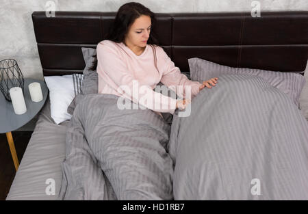 Junge Frau aufwachen ihr beleidigt Freund, der sich mit einer Decke bedeckt, während im Bett liegend und tragenden Pyjamas, in der Nähe von Nachttisch mit cand Stockfoto