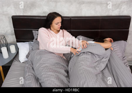 Junge Frau aufwachen ihr beleidigt Mann, der sich mit einer Decke bedeckt, während im Bett liegend und tragenden Pyjamas, in der Nähe von Nachttisch mit Kerze Stockfoto