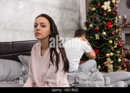 Junges Paar sitzen Rücken an Rücken auf Bett und ignorieren einander im Schlafzimmer im loft-Stil mit grauen Farben und Weihnachtsbaum mit präsentiert Stockfoto