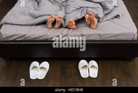 Männliche und weibliche Füße, Hand des Mannes, seine Füße unter grauen Decke in modernes stilvolles Bett in der Nähe von zwei Paaren von weißen Hausschuhe kratzen Stockfoto