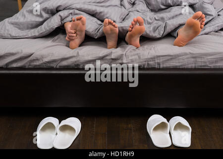 Männliche und weibliche Füße, zarte Frauenhand kratzen ihre Füße unter grauen Decke in modernes stilvolles Bett in der Nähe von zwei Paaren von weißen Hausschuhe Stockfoto