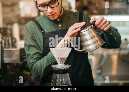 Kellner mit schwarzer Schürze und Brillen gießt heißes abgekochtes Wasser im Kaffee Topf stehen am Tresen. Stockfoto
