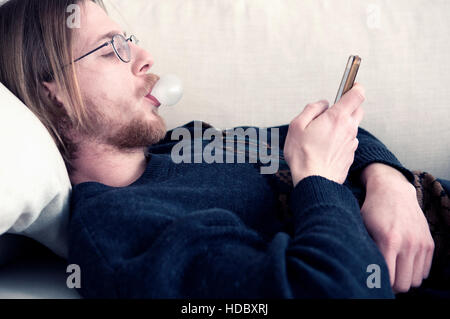 junge rothaarige bärtiger Mann mit bubblegum liegend auf der Couch und SMS auf seinem Handy Stockfoto