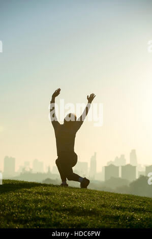 Silhouette eines Mannes stretching auf grasbewachsenen oben Primrose Hill vor einem nebligen goldenen Sonnenaufgang Blick auf die Skyline von London Stockfoto