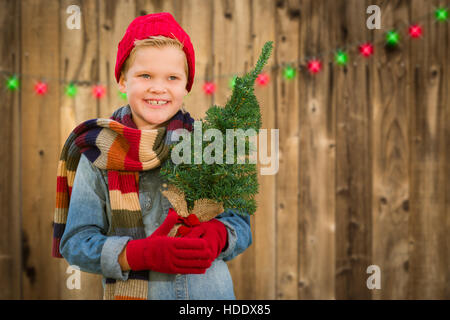 Glückliche junge tragen, Schal und Handschuhe halten Weihnachtsbaum auf einem Holzzaun-Hintergrund. Stockfoto