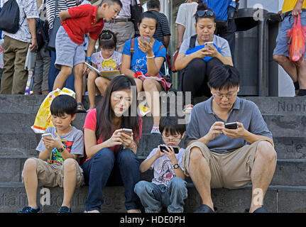 Eine asiatische Familie sitzen auf den Stufen in der Stadtbibliothek in Downtown Flushing, Queens, New York, alle mit ihren Handys. Stockfoto