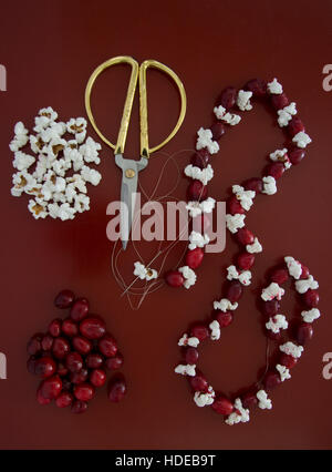 Eine teilweise gemacht Weihnachtsgirlande mit mehr Popcorn und frischen Preiselbeeren, zusammen mit Gold behandelt Schere auf einem roten Tisch Stockfoto