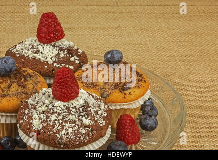 4 Muffins mit geriebener Schokolade bestreut und garniert mit frischen Himbeeren und Heidelbeeren auf einer Glasplatte und der ganze Bob auf der Tischdecke Stockfoto