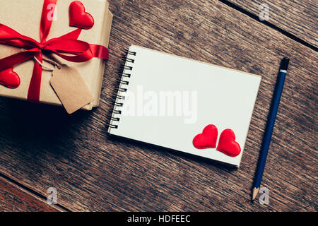 Draufsicht (oben) Geschenk-Box und rotes Band und rotes Herz mit Notebook zum Valentinstag auf Tisch aus Holz. Valentine Hintergrund. Stockfoto