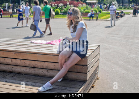 Blonde Frau trägt Sonnenbrille und Jeans Minirock, sitzt auf einer Holzbank. Gorki Park, Moskau Stockfoto