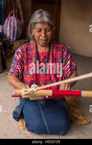 Eine ältere, grauhaarige Frau Maya webt Stoff auf einem Backstrap Loom kniend auf dem Boden ihres Hauses in Santa Catarina Palopo, Guatemala. Stockfoto