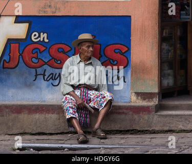 Ein älterer Maya-Mann in traditioneller Kleidung von San Pedro la Laguna, Guatemala, sitzt durch eine gepflasterte Straße vor eine bunt bemalte Wand, die sagen Stockfoto