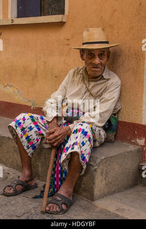 93 Jahre alte Maya-Mann in traditioneller Kleidung befindet sich auf Straße in San Pedro la Laguna, Guatemala. Sitzen auf dem Bordstein mit seinem Stock vor einem Pastell pa Stockfoto