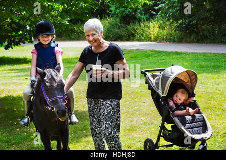 Frau, Baby im Kinderwagen und Blond Mädchen sitzen auf einem Pony reiten-Hut trägt. Stockfoto