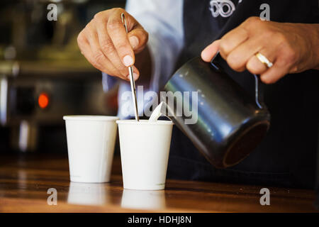 Nahaufnahme von heißer Milch in einen Pappbecher mit einer Kanne gegossen wird. Stockfoto