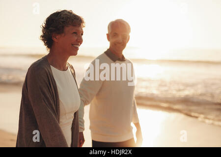 Lächelnde senior Frau mit ihrem Ehemann zu Fuß am Strand bei Sonnenuntergang. Älteres Paar zu Fuß entlang der Küste. Stockfoto