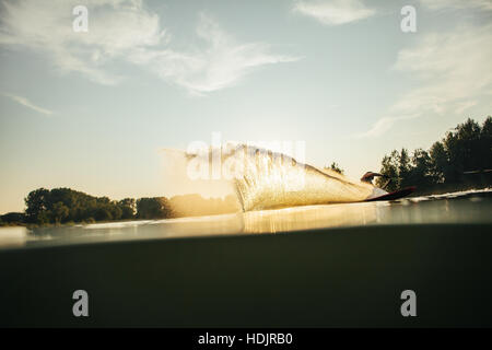 Wasserschifahrer bewegt sich schnell in Spritzwasser bei Sonnenuntergang. Mann Wakeboarden auf einem See Stockfoto