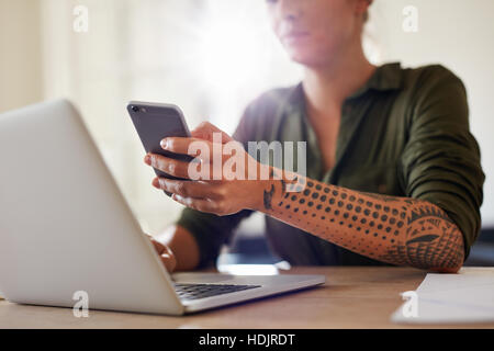 Aufnahme der jungen Frau mit Handy während der Arbeit am Laptop. Smartphone in der Hand einer Frau im Mittelpunkt. Stockfoto
