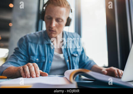 Junger Mann sitzt am Tisch mit Büchern und Laptop zum Auffinden von Informationen. Junge männliche Student Studium in Bibliothek Buch zu lesen. Stockfoto